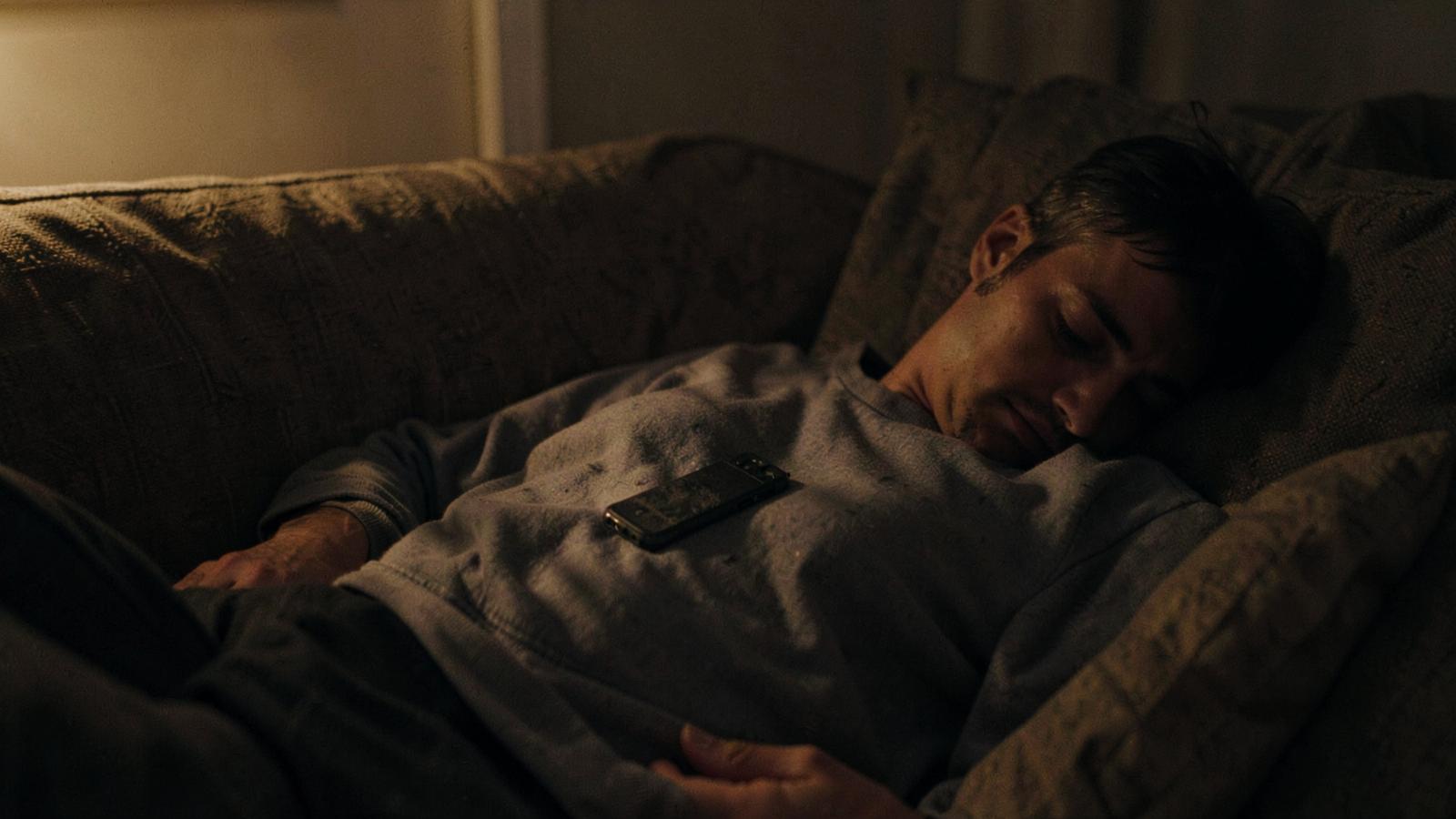A person lying on a sofa in dim evening light with a phone face down on their chest