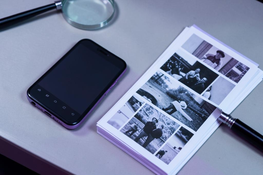 A phone face up on a desk beside a magnifying glass and a stack of printed photographs